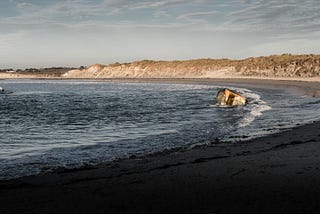 Photograph of a small concrete bunker on a beach being lapped by waves.