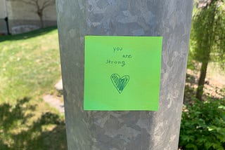 Lime green sticky note on a light post saying, “You are strong.”