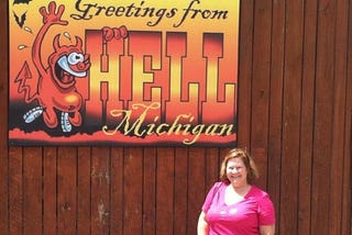 woman stands in front of a sign which says “greetings from Hell Michigan. politics, inauguration, hell