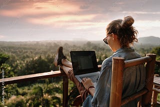 Woman sitting on chair on deck overlooking wilderness. She has feet on rail while working on laptop.