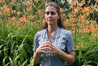 A woman kneeling with her fingertips touching. Her thumbs are against her chest and her elbows are relaxed against her body. She’s wearing a black and white vertically striped shirt in front of a patch of orange flowers.