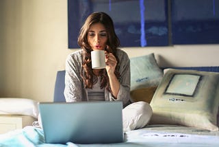 Woman in a grey sweater sitting on a couch drinking coffee and using her laptop