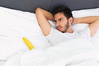 A young man in a white teeshirt looks perplexed at he stares at a banana emerging from behind the sheets