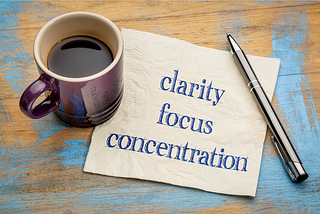 A cofffe mug, a napkin with the words clarity, focus and concentration written with a pen laying on the side
