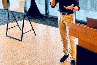 Orlando T White presenting at Stanford University Oak Room. Orlando is smiling, holding a microphone, and wearing a black shirt and khakis.