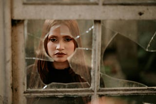 A sad woman staring through a broken window.