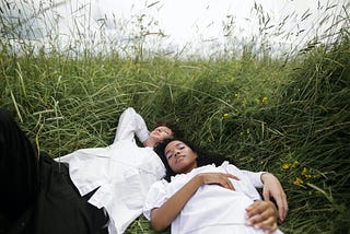 Two individuals laying in a bed of grass together with their eyes closed wearing white T-shirts