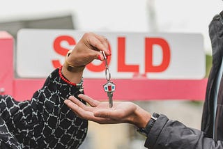 A person handing over a key in front of a “Sold” sign