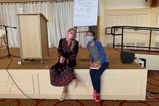 A white blonde haired women with wavy hair wearing a pink mask and wearing a red and blue bohemian style dress is smiling directly at the camera and is wearing pink sneakers on the left. A brown haired women is wearing a blue mask wearing a gray long sleeved t-shirt with blue sleeves, dark blue leggings and one pink sock on the left foot and a red sock on the right foot. They are both sitting on a stage that the floor is brown, with a tan podium in the back.