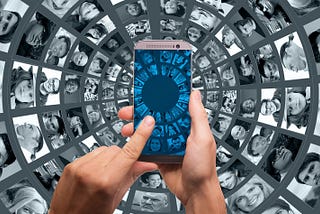 A photo of hands holding a cell phone against a backdrop of multiple screens with people’s faces in them.