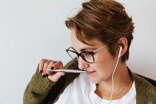 Woman with green sweater, wearing spectacles, and ear buds. Holding a pen to her mouth and gently smiling, gaze down.