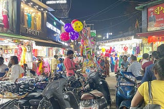Motorcycles are parked in the center of this picture. Someone’s selling socks off one of the motorcycles. A toy seller sells balloons and toys. Clothes shops display their wares, There are plenty of people in this picture. Some people are walking around with their motorcycle helmets on.