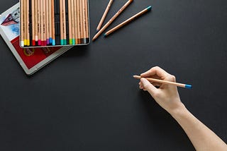 An image of a box of colored pencils on a black background. A hand holds one of the pencils near the middle.