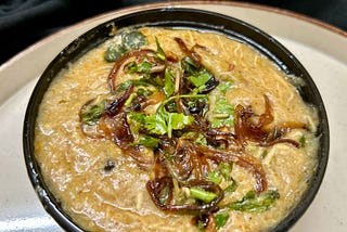 On a white plate, a black bowl holds a portion of brown-colored stew with shredded meat, garnished with caramelized onions, coriander leaves, and mint leaves.