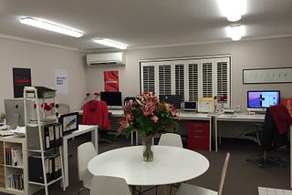 An office at night. Four white desks, topped with mac computers, red accents around the place. A round white table topped with red flowers sits in the foreground.