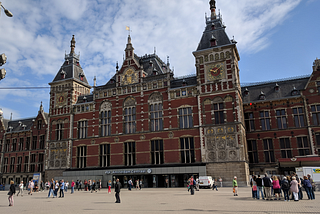 Amsterdam’s main train station is an ornate red building with three sloped top towers. Two clock towers on the left and right, and the center tower is wider. People are walking or standing around in the plaza in front.