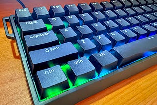 A closeup of the left half of an Aula Win68 gaming keyboard, with the lighting set to blue and green under the black keys. The upper left of the keyboard body has a silly hook on it for an included strap, which isn’t installed.