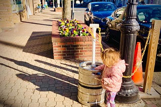 image shows a scraggly haired toddler in a pink coat on a brick sidewalk, playing with a metal keg that is next to a black lamppost. In the background, there is a brick planter with colorful flowers