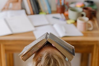 A person is sitting at a desk with books scattered on top. Their head is tipped back with an open book covering their face.