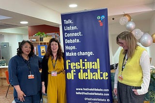 The three researchers standing next to the Festival of Debate poster, which is blue with yellow writing. Yinka is wearing blue and Christina & Louise are wearing yellow.