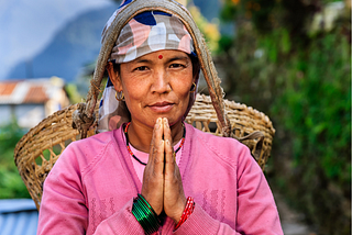 A Nepali woman folding her hands in front of her chest. She is wearing a basket on her back with the string going across her forehead
