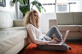 A woman sits on the floor of her living room typing on her laptop.