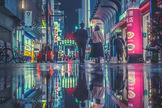 Two people walk down a colorfully lit city street. It appears as though it has just rained. The lights of the city reflect off of the glassy ground, giving the image a magical quality.