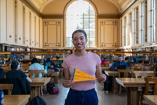 Zoë poses for a photo in Doe Library on UC Berkeley’s campus while holding a pennant that states “I am Barbadian-German-American“
