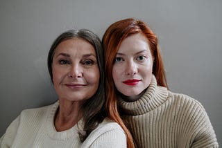 An older woman with dark skin and grey, long hair in a white sweater has her head next to a young woman with light skin and red, long hair.