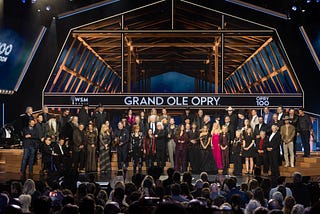 A large group of Grand Ole Opry members from multiple generations, including Bill Anderson, Connie Smith, Randy Travis, Reba McEntire, Carrie Underwood, and Carly Pearce, stand together on stage at the Opry House during the 100th Anniversary Celebration in 2025, beneath the Grand Ole Opry sign and wooden stage beams, honoring a century of country music tradition.