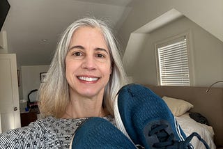 Woman in a bedroom smiling and holding up a pair of blue sneakers