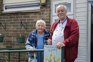 Photo of an older couple, Wilf and Carol as they stand outside a home. They hold hands and smile slightly at the camera.