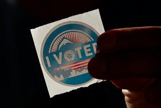 Image of a person holding an “I Voted” sticker. Photo via Unsplash, courtesy of Manny Becerra (@mannyb).