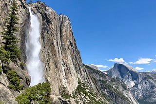 The raging water of Yosemite Falls tumbles over the sheer granite cliff. Half Dome towers in the background.