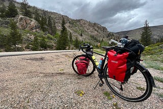 The author’s bicycle, on the Cameron Pass over the Rocky Mountains. It has two red pannier bags over the rear wheels and two red pannier bags over the front wheels; in the background are mountains and a cloudy sky