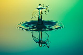 Droplet of water mirrored perfectly on a clear water surface with a yellow green gradient background.