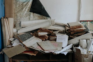 A mountain of ageing papers and files on a desk in a poorly lit room. By Bezya Kaplan