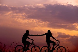 A couple bicycling together in the sunset, the woman is reaching her arm out to the man.