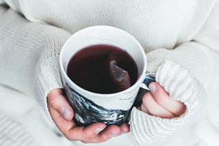 A person in a white sweater holding a dark cup of tea. The teabag is still in the water.