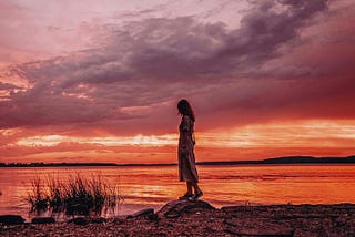 A woman stands alone by a lake at sunset, silhouetted against a dramatic sky of purples and reds. Her posture is contemplative, evoking solitude, surrender, and the quiet beauty of letting go.