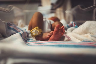 A newborn rests on a hospital bassinet covered with a white blanket featuring pink and blue stripes. The baby’s legs are slightly bent, and the focus is on the tiny feet. One ankle has a yellow monitor band, and the other has a pink hospital ID band. The lighting is soft and warm, highlighting the delicate skin and the peaceful atmosphere of the hospital setting.