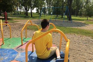 A young boy sitting with his back to the camera on a merry-go-round at a park. There are trees in the background.