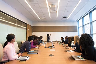 In a conference room, a diverse group of employees with laptops in front of them are facing the whiteboard seeming to listen to another employee speak as she write on the whiteboard.