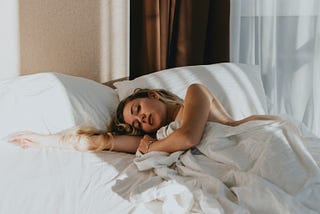 Blond woman with bedroom eyes, draped in a white sheet and lying on white pillows, stretching an arm toward the empty side of the mattress