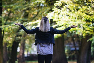 A white woman with short brown hair and a blonde balayage wearing a dark jack and a white shirt looks straight up shrugging her arms at the sky as if to ask the very heavens Why.