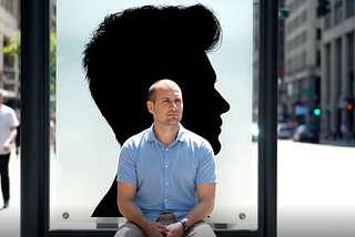A bald man sitting at a bus stop in front of a large advertisement showing a silhouette of a man with a full head of hair.