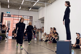 Three dancers stand in a gallery space with white walls. They are all wearing black dresses. One stands upon a grey cube, elevated above the others. An audience watches on.