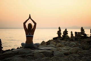 A woman in sports clothing meditating on a jetty near the open ocean. There is high contrast between the black rocks of the jetty and the brightness of the sea and sky.