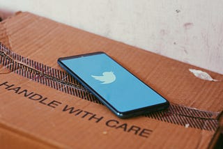 Smartphone with the Twitter app started on a cardboard box with the words, “Handle With Care.”