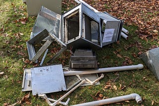 A photograph showing parts of an old HVAC system spread out on a lawn. There are fallen brown leaves on the grass, especially at the top right of the photo.